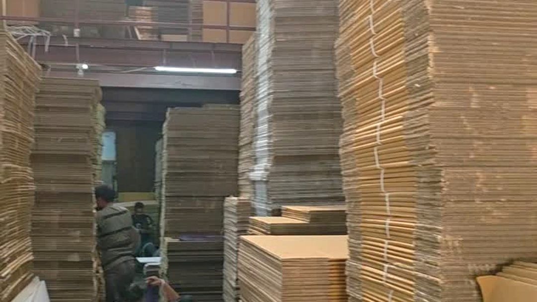 Inside the Next Door Packaging godown at Bhumi World, Bhiwandi — stacks of flat corrugated board and finished cartons, with two of the team working between the rows.
