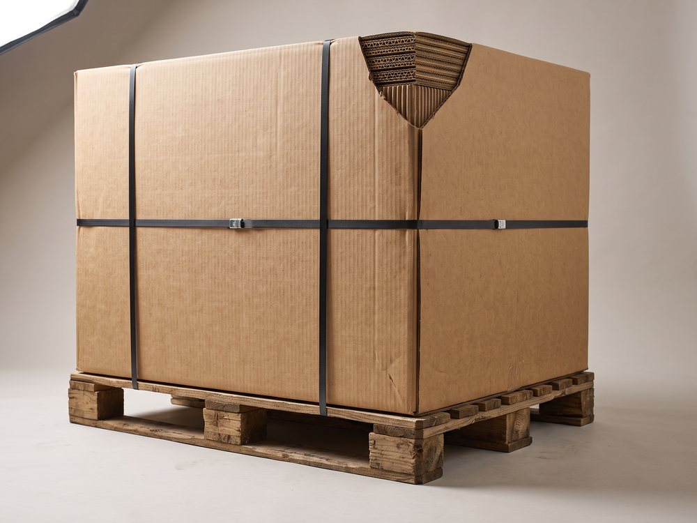 A large double-wall corrugated carton strapped onto a wooden pallet, with the two flute layers visible at the cut corner.